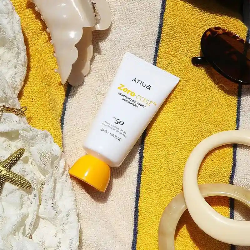 Tubo do protetor solar Anua Zero-Cast sobre toalha de praia, com óculos de sol e acessórios
