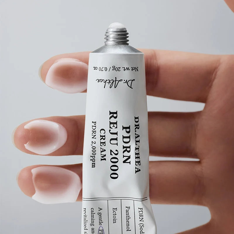 Mão a segurar o tubo de 20g com porções de creme branco aplicadas nas pontas dos dedos