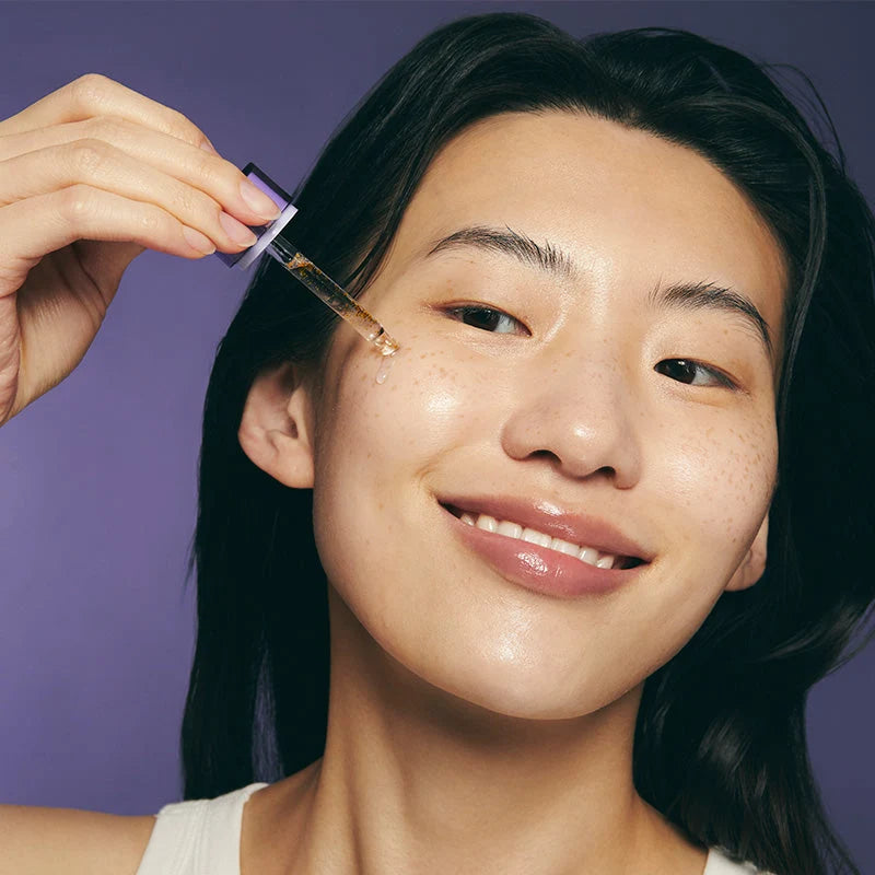 Mulher de cabelo escuro a aplicar uma gota de sérum na bochecha com o conta-gotas, sobre fundo roxo
