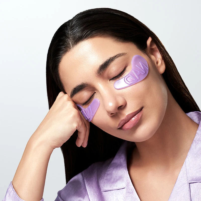 Modelo com os olhos fechados e um patch aplicado sob cada olho, vestindo roupão roxo.